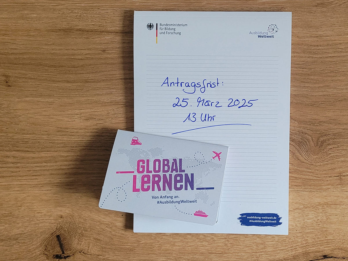 Notizblock auf dem per Hand geschrieben steht: "Antragsfrist 25. März 2025, 13 Uhr". Im unteren linken Drittel liegt ein kleiner Block mit Haftnotizen, auf dem Umschlag steht "Global Lernen. Von Anfang an. #AusbildungWeltweit".