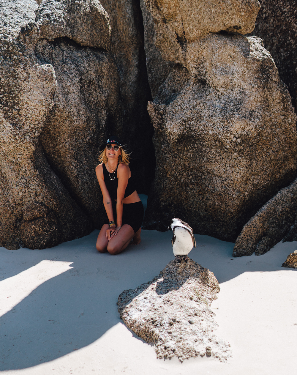 Ein Pinguin im Vordergrund auf einem Stein am Strand, im Hintergrund Franzi im Sand knieend
