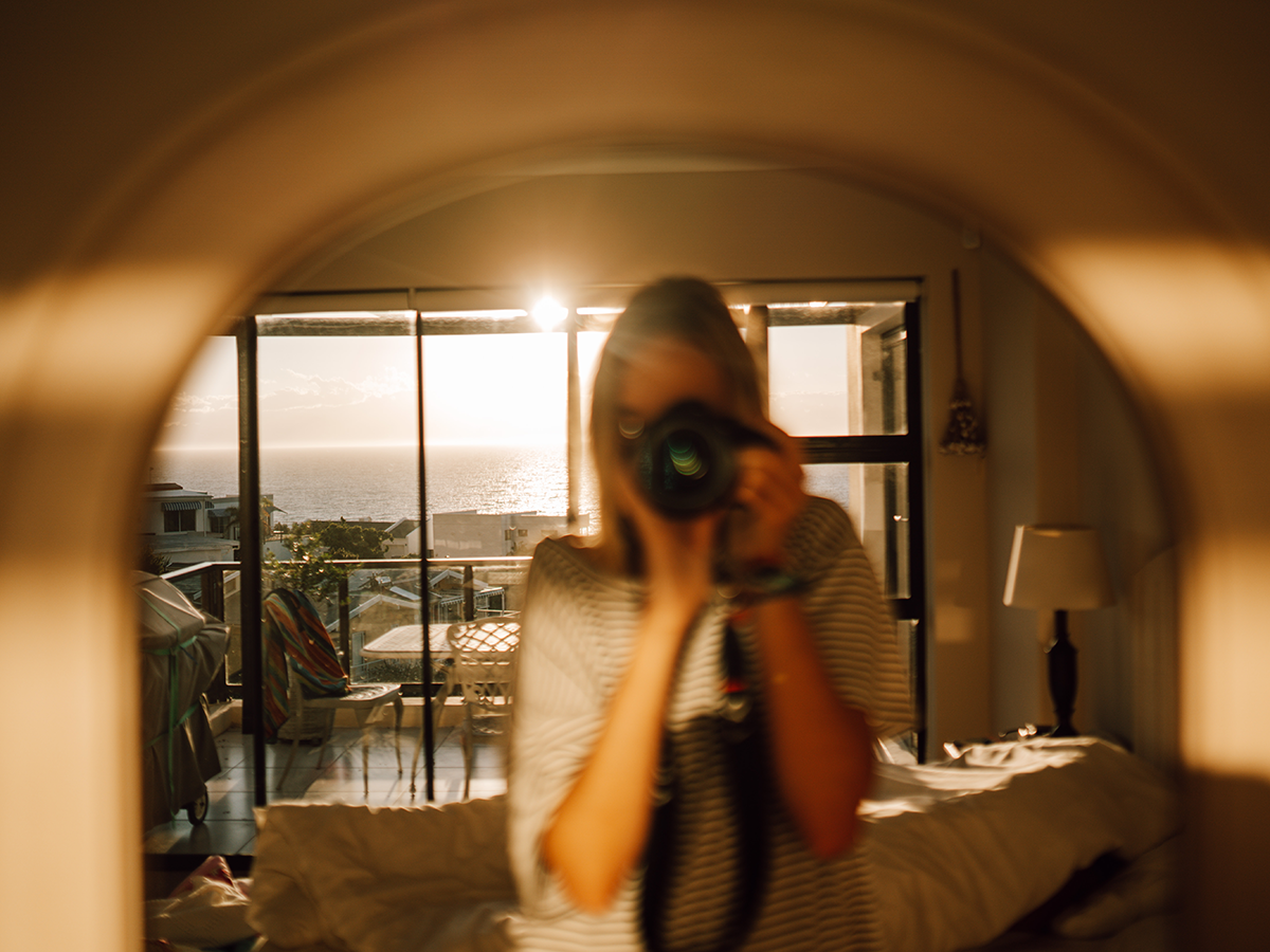 Selbstportrait mit Kamera vor Gesicht im Spiegel mit Scharfstellung auf das Zimmer und den Ausblick im Hintergrund