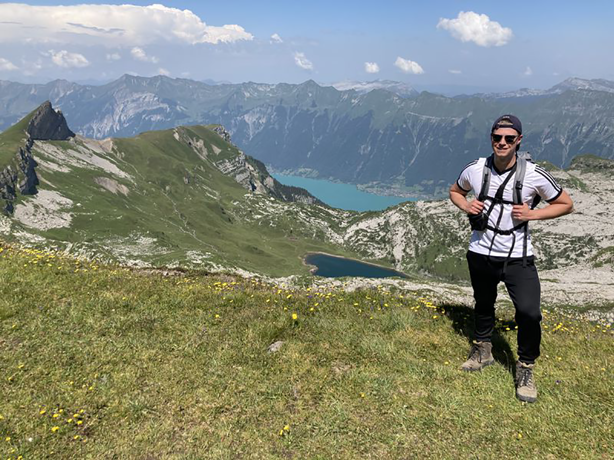 Azubi Markus steht vor einer Aussicht auf einen See in den Schweizer Bergen